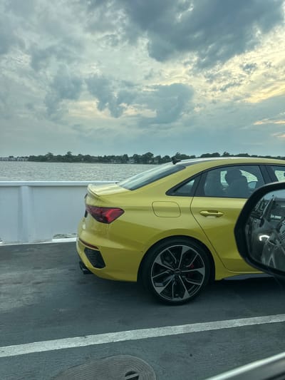 A bright yellow Audi sedan is parked on a ferry, viewed from the side and rear. The car's taillights and rear spoiler are visible. In the foreground, the ferry deck is dark asphalt with white painted lines. Beyond the car, a body of water is visible with a distant shoreline dotted with houses and trees under a cloudy sky. The reflection of another vehicle in the Audi's side mirror shows its headlights. The scene appears to be located in or near the Village of Greenport, United States of America.