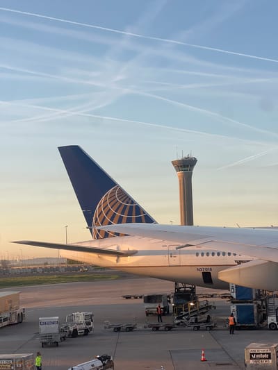 The image shows a United Airlines airplane, specifically N37018, at Roissy-Charles de Gaulle Airport (CDG) in Roissy-en-France, France. The tail of the plane is visible, displaying the United globe logo. A control tower is seen in the background, as well as contrails in the sky. Ground crew and equipment are visible around the plane, including cargo containers with labels like "Munilode" and "AKE 20507 UA." One ground worker is wearing a reflective vest.