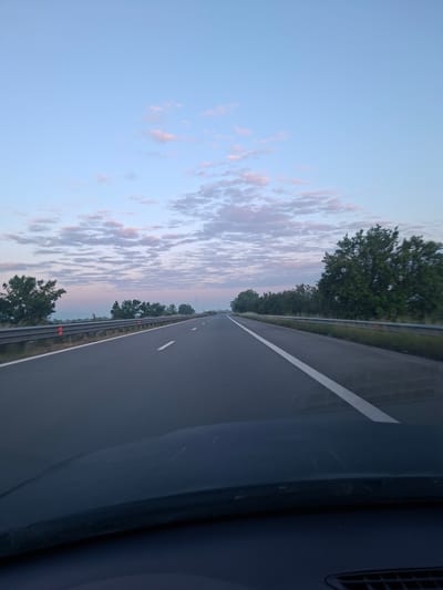 The image shows a multi-lane highway under a sky with light clouds at sunset. The view is from inside a vehicle, looking out across the road. A median with trees runs alongside the highway. There is no visible indication that the scene is in Shishmantsi, Bulgaria. There are no people visible.