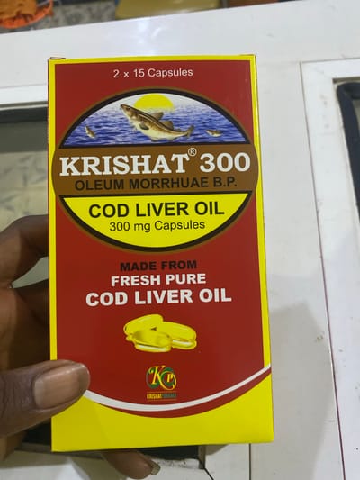 A person's hand holds a yellow and red box of "Krishat 300 Cod Liver Oil". The box has a picture of two fish in the ocean with a sun. Text on the box reads "2 x 15 Capsules", "Krishat 300 Oleum Morrhuae B.P.", "Cod Liver Oil", "300 mg Capsules", "Made From Fresh Pure Cod Liver Oil", and a logo that says "Krishat Pharma". The image appears to be taken indoors, with a white surface visible in the background. The location is not specified in the image.