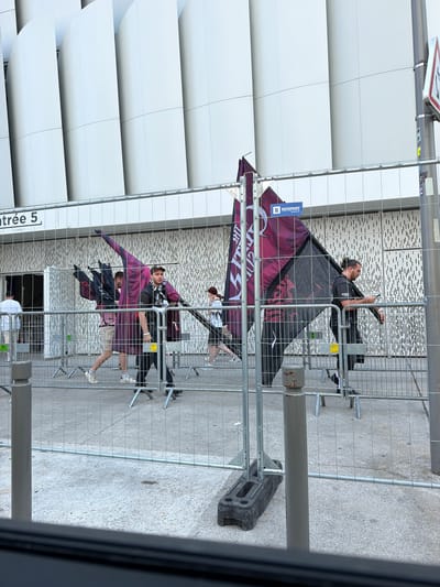 This media file depicts an outdoor scene in Nanterre, France, likely during the daytime under fair weather conditions, judging by the lighting. The primary focus is on several individuals situated behind a metal safety fence. The fence runs diagonally across the frame, creating a sense of depth.
In the foreground, the fence is composed of silver metal poles and wire mesh. Beyond the fence, a modern building with white, curved facade elements is visible. The ground appears to be paved with light-colored, textured material, possibly concrete or stone.
The individuals are engaged in various activities. One person, wearing a dark t-shirt and with their hair tied back, is holding a mobile phone. Another individual, wearing a purple outfit and shorts, is standing with their arms outstretched, appearing to be posing or participating in an activity. Several other people are partially visible or out of focus in the background. Some are holding large, dark-colored flags with what appear to be red or maroon designs.
Notable details include a "RÉMPART" sign attached to one of the flags, indicating a brand or company. There is also text on the building that reads "entrée 5" and "Gate," suggesting this is an entrance area. The overall atmosphere seems casual and possibly related to an event or gathering. The subjects' expressions are not clearly discernible due to the image quality and distance.