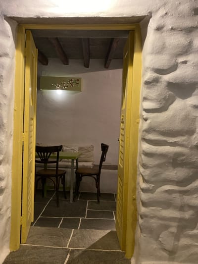 The image displays an interior view, looking through an open double doorway into a compact room, identified as being in Apollonia, Greece. The doorway, framed by bright yellow, louvered panels, is set within thick, whitewashed, textured stone walls. The floor, both in the immediate foreground and extending into the room, is composed of large, irregularly cut dark grey flagstones with light grout lines. Inside the room, the walls are smooth and light-colored, contrasting with exposed dark wooden beams on the ceiling. A light-toned, horizontal wall-mounted fixture, adorned with a branch-like motif featuring small red elements, is positioned high on the far wall, casting a downward-directed light. Directly below this, a square table with a light green tabletop and dark legs is centered. Two dark brown, bentwood-style chairs are placed at the table, one on either side. Along the back wall, behind the table, a low, light-colored cushioned bench or banquette is visible. No people are present in the scene.