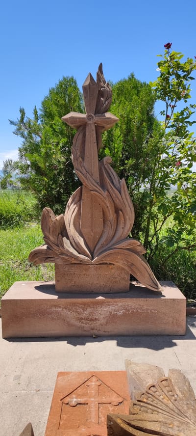 The image depicts a stone sculpture in the city of Araks, Armenia. The sculpture is of a cross emerging from stylized flames or petals, carved from reddish-brown stone. It stands on a rectangular stone base, which is itself on a paved area. Behind the sculpture are lush green trees and grass, under a bright blue sky. The scene is set outdoors, with natural lighting. The sculpture appears to be a monument or memorial, with inscriptions on the base, though these are not clearly legible in the image. In the foreground, there are other stone fragments, one of which has a carved cross. The overall impression is one of solemnity and natural beauty.
