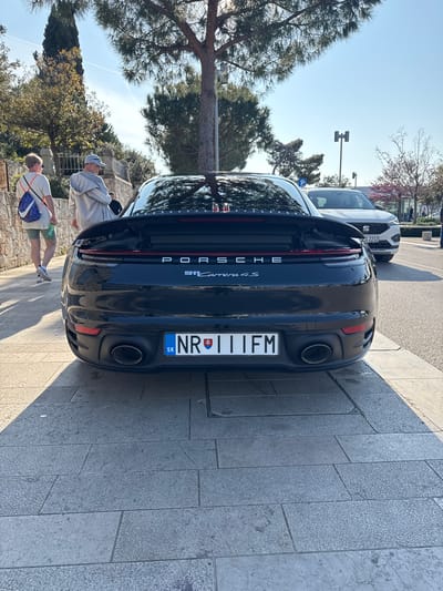 The image captures the rear view of a dark-colored Porsche Carrera 4S, displaying a Slovakian license plate, "NR 111FM." The car is parked on a stone-paved surface that suggests a pedestrian area. In the background, blurred figures of two people are walking near the car. The setting appears to be a sunny day near trees and stone architecture. Given the distinctive stone pavement and architectural style, it is highly likely the location is in the city of Grad Rovinj, Croatia. Another car is seen parked in front of the Porsche.