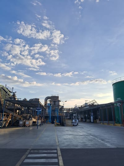 The image depicts an industrial scene, likely within or near a manufacturing facility. The location is identified as Chimbote, Peru.
The frame is split between a bright, cloudy sky and a concrete or asphalt roadway. Heavy industrial infrastructure lines the right side of the road. This includes tanks, pipes, and metal structures. A white truck is parked along the road, closer to the background.
The sky has varying cloud formations against a blue backdrop. The roadway features yellow painted lines and a pedestrian crosswalk.