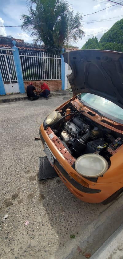 This image captures an outdoor scene in El Papelón, Venezuela, featuring an orange or bronze-colored car undergoing repairs. The car is parked on the side of an asphalt road, with its hood wide open, revealing the engine compartment. The engine has a cover with "DODGE" visible on it, and a black aftermarket air filter is prominent. One of the car's headlights appears hazy, while the other is either removed or obscured. The front bumper shows signs of wear, with chipped paint. A license plate, partially visible, reads "NBN-27MG." A black rubber mat is placed on the ground in front of the car, and a jack stand is partially visible, supporting the vehicle.
Two men are engaged in activity on the concrete sidewalk adjacent to the car. One man, dressed in a red shirt and blue pants, is kneeling. The second man, wearing a black shirt and black pants, is also kneeling, facing the first, and both appear to be focused on a task on the ground, likely related to the car's maintenance.
In the background, a residential house with a white security gate, blue trim, and a tiled roof is visible. A large palm tree stands prominently, along with other dense green foliage, against a sky with scattered clouds. The bright daylight and clear shadows suggest the scene takes place during the midday or early afternoon. The overall impression is one of practical work being carried out on a vehicle in a residential street setting.