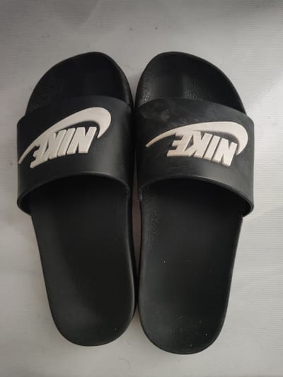 This is a top-down, eye-level, full-shot, medium-full photograph of a pair of black Nike slide sandals. The sandals are placed side-by-side on a white, textured surface. Each sandal features a broad, black strap with a white, raised Nike logo. The lighting is even and diffused, casting no significant shadows. The background is a plain white surface with a subtle ribbed texture. The image's quality is clear, with no visible dust or debris on the sandals. The scene is static and unpopulated, suggesting an indoor setting. The location of Snezhinsk, Russia, is not visually represented in the image; this information is external to the visual content.