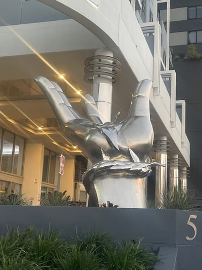 A large, polished silver sculpture of a hand is prominently displayed in front of a modern building in Brisbane, Australia. The sculpture depicts a hand with a laurel wreath wrapped around its base. The building features a white, multi-story facade with balconies and large windows, and a dark gray structure rises behind it. The sculpture is positioned against a backdrop of urban architecture, with a gray wall in the foreground adorned with the number "5". Lush green plants are visible both in front of and behind the wall, suggesting a well-maintained urban landscape. The lighting indicates it is daytime, with sunlight casting shadows and highlights on the metallic sculpture and the building. A red and white "Do Not Enter" sign is visible on a pole near the sculpture.