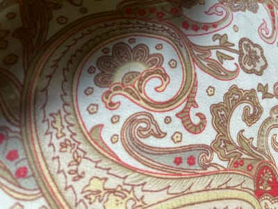 The image displays a close-up view of a fabric with a traditional paisley pattern. The pattern features swirling motifs in shades of gold, brown, and red against a light-colored background, possibly white or cream. Small floral accents in red are scattered throughout the design. The texture of the fabric is visible, showing a woven material. There is no identifiable location in the city of Orozbekov, Kyrgyzstan, as the image is solely focused on the textile design.
