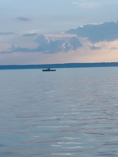 A boat with a person on board is seen in the distance on a large body of water under a cloudy sky. The boat is dark-colored and appears to be a motorboat, with a single person standing in it. The water is calm with gentle ripples, reflecting the light blue and white of the sky and clouds. A dark, tree-lined horizon is visible in the background. The overall scene is serene and suggests a quiet day on the water. This scene could be located in the city of Галево, Russia, given the presence of a large body of water.
