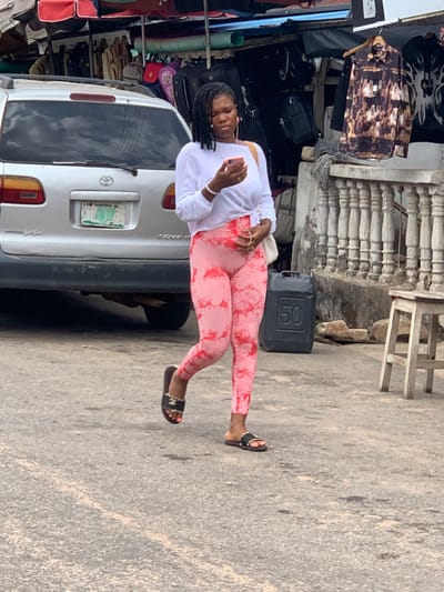 A woman in pink leggings and a white shirt stands in front of a silver Toyota van. The van has a Lagos, Nigeria license plate. She is wearing black sandals and holding a phone. Behind the van, there are various bags and other items for sale, suggesting an outdoor market. A black jerrycan is next to a concrete structure, with a wooden stool nearby. The scene appears to be set on a street.