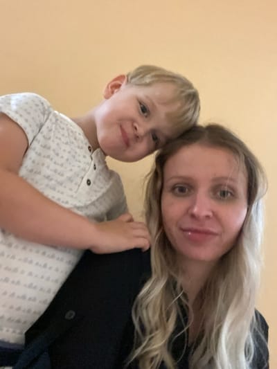 In this close-up, low-angle selfie, a young child with blond hair leans over the shoulder of an adult woman, resting his head on her head. Both individuals are looking towards the camera. The child wears a white t-shirt with a subtle grey pattern. The woman has blond hair and is wearing a dark top. The background is a plain, light orange wall, characteristic of an interior setting. The image quality is somewhat grainy and lacks sharp detail, suggesting it was taken with a mobile device. The overall impression is an informal, candid moment captured between a mother and her child. No specific details of Shiryaevo, Russia, are discernible from the provided image.