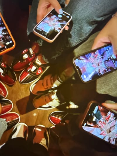 This media file shows a group of people, or at least their hands and feet, gathered around a reflective floor. Several individuals are holding up their smartphones, which display images of what appear to be bowling shoes arranged in a circular pattern. The shoes themselves are a mix of red, black, and white, with some featuring a circular logo that resembles the letter 'G' within a circle. The hands holding the phones have manicured nails, with one hand showing light blue nail polish. The overall scene suggests a casual gathering, possibly at a bowling alley, given the presence of bowling shoes and the reflective floor commonly found in such establishments. The lighting appears to be artificial, creating reflections on the floor that add to the visual effect. While the exact time of day or weather is not discernible, the indoor setting and the focus on the activity imply a planned social event. The emotion conveyed is one of shared experience and amusement, as evidenced by the act of taking pictures of each other's footwear. The location context of Pori, Finland, provides a specific geographical setting for this social gathering. There is no visible text in the image, other than the graphical elements on the bowling shoes and the phone screens.