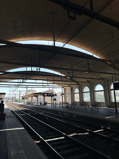 This is a photograph taken at a train station in Nîmes, France. The image captures the underside of a curved concrete structure that shelters a train platform and tracks. Sunlight streams through the gaps in the structure, casting dramatic shadows and illuminating parts of the platform and the railway lines. Arched windows are visible on a building behind the platform, suggesting an older architectural style.

The scene is likely set during the daytime, given the strong sunlight. The weather appears clear. On the platform, there are signs, benches, and what appear to be parts of train station infrastructure like overhead wires and poles. The railway tracks stretch into the distance, with ballast and sleepers clearly visible. In the foreground, a few pieces of luggage can be seen on the platform, hinting at people waiting for trains. The overall mood of the image is one of quiet anticipation, with the play of light and shadow adding a sense of drama and depth to the urban environment.

There are some signs visible in the image. One sign near the tracks has numbers "26" and "25" on it. Another sign higher up appears to be a timetable or route information, with some text in French, including "SNCF".