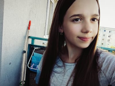 A young person with long, dark hair and fair skin is taking a selfie on a balcony. They are wearing a light gray t-shirt and a small hoop earring in their left ear. The person's face is partially obscured by their hair, but their eyes, nose, and mouth are visible. The background shows a concrete wall, a window, a green railing, a drying rack with folded blue laundry, a broom with a red handle, and a light-colored apartment building with several windows. The scene appears to be outdoors, with natural light illuminating the area. The specific location of the balcony is in Radzymin, Poland.