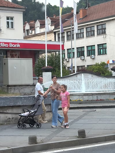 In Konjic, Bosnia and Herzegovina, a family is walking on a paved street in front of a building that houses "Diko Bank." A man in a white shirt and tan pants pushes a gray stroller with a baby inside. He is walking next to a woman wearing a brown top and blue jeans, who is holding an ice cream cone and walking with a young girl in a pink t-shirt and peach shorts. In the background, a fountain sprays water, and trees line the street behind the buildings. The sky is overcast.