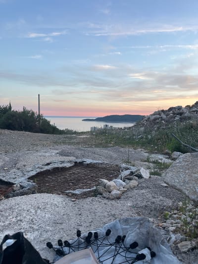 The image depicts a scenic overlook in the city of Čučuci, Montenegro. The foreground is a rugged, undeveloped area strewn with rocks and debris, including a discarded mattress spring, concrete fragments, and a black plastic bag. A white plastic sheet is also visible, partially covering what appears to be a string of black electrical components. In the mid-ground, a patch of soil is enclosed by rocks, suggesting a potential garden or planting area. Beyond this, a body of calm water, likely the Adriatic Sea, stretches towards a distant coastline dotted with buildings. The horizon is painted with the soft hues of a sunset, with streaks of pink and orange clouds against a pale blue sky. A tall, slender industrial chimney stands out against the tree line on the left, while other smaller structures are visible on the distant shore. The overall impression is one of an urban fringe meeting natural beauty at dusk, with elements of construction and neglect in the foreground.