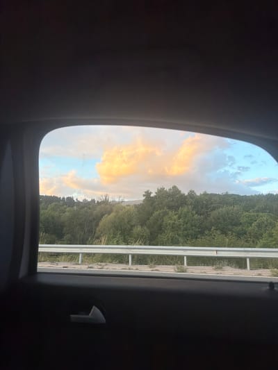 The image captures a view from inside a vehicle looking out of a rear window at a rural landscape. The window frame is dark and partially obstructs the view. In the distance, a dense forest covers rolling hills, with trees of varying shades of green. A section of a guardrail, appearing to be made of metal, runs horizontally across the middle ground. Beyond the guardrail, there is a glimpse of what seems to be a road or embankment with some vegetation. The sky above is partially filled with clouds; on the left, large, puffy clouds are illuminated with a warm, golden-orange light, suggesting either sunrise or sunset. On the right, the sky transitions to a pale blue with scattered white clouds. The image was taken in Studena, Bulgaria.