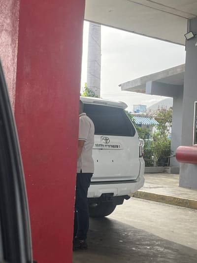A man in a white polo shirt and dark pants is partially obscured by a red pillar, standing next to the rear of a white Toyota 4Runner in what appears to be a service area or parking lot in San Antonio, Venezuela. The vehicle has a license plate attached to the rear bumper. The environment suggests an outdoor setting with overhead structures and vegetation visible in the background. The lighting indicates it is daytime.