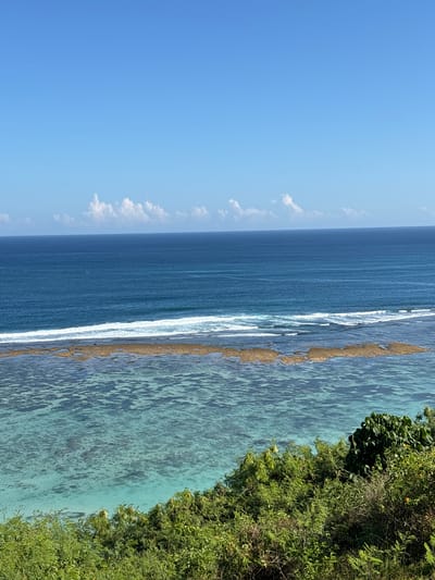 The image depicts a coastal view, likely taken from an elevated position near Benoa, Indonesia, with a bright blue sky occupying the upper left portion of the frame. Thin, white clouds are scattered near the horizon. The ocean transitions from a darker blue further out to a lighter, turquoise color closer to the shore, indicating shallow water. A line of white surf breaks along what appears to be a coral reef or sandy bottom, separating the deeper blue water from the shallower turquoise area. Lush green vegetation, possibly a cliffside or hillside, occupies the right side of the frame. There are no visible people or animals in the scene. The clear visibility suggests a sunny day with good weather conditions.
