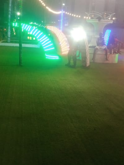 The image captures a nighttime scene in a public park or square within Majengo ward, Kenya. The ground in the foreground and midground consists of artificial green turf. A thin pole, possibly a tree or light standard, stands vertically in the left foreground, illuminated with green light. In the midground, a series of illuminated archway structures extends horizontally. The leftmost visible arch emits green light, the next displays blue and white light, and a third to the right shows amber/orange and white light.

An intense, bright white light source is positioned centrally in the midground, obscuring the upper torso of an individual standing in front of it. This person's lower body is visible, clad in dark trousers and shoes. To the right, two or three individuals are seated on a white, ornate concrete structure, possibly a bench or decorative element. One individual is clearly visible, wearing a plaid shirt, light-colored shorts, and dark shoes, with their legs dangling. Above, a string of warm white decorative lights stretches across the upper portion of the frame. The background features indistinct architectural shapes and faint outlines of additional individuals.