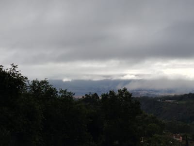 The image depicts a cloudy, overcast sky over a green, hilly landscape. In the foreground, dense foliage, likely trees, obscures much of the ground. Beyond the immediate trees, the terrain slopes downwards, revealing a valley with scattered buildings and structures, suggesting a semi-rural or suburban environment. In the distance, more hills and a hazy, indistinct settlement can be seen, under a thick layer of low-hanging clouds that blend with the distant landscape. The overall impression is a muted, somber atmosphere due to the heavy cloud cover. The scene is consistent with the natural topography and vegetation found in the outskirts of Arucas, Gran Canaria, Spain, where hilly terrain meets more populated areas under varying weather conditions.