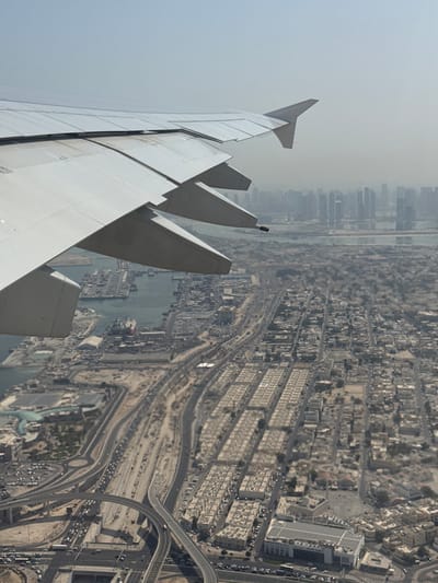 The image provides an aerial view of a coastal city, likely Deira in the United Arab Emirates, from the perspective of an airplane window. The airplane wing occupies the upper left quadrant of the frame. Below the wing, a sprawling urban landscape unfolds, characterized by dense residential areas with uniformly constructed, low-rise buildings arranged in grid patterns. Numerous roads and highways weave through the city, some elevated with complex interchanges, suggesting a developed infrastructure. Traffic, consisting of numerous vehicles, is visible on these roads, indicating a busy urban environment.
Further into the background, a port or harbor area is discernible, with numerous vessels docked and cargo containers organized on the land. Beyond the port, the distinctive skyline of a modern city emerges, with numerous tall skyscrapers piercing the hazy atmosphere. The overall environment appears somewhat arid, typical of a desert climate, with muted earth tones dominating the landscape, interspersed with the blues of the water and the grays and whites of the urban structures. The sky is a pale, hazy blue, suggesting either a dry, dusty atmosphere or a distant view. There are no discernible people or distinct actions, the scene captures a static, expansive view of urban development and activity from a high vantage point.