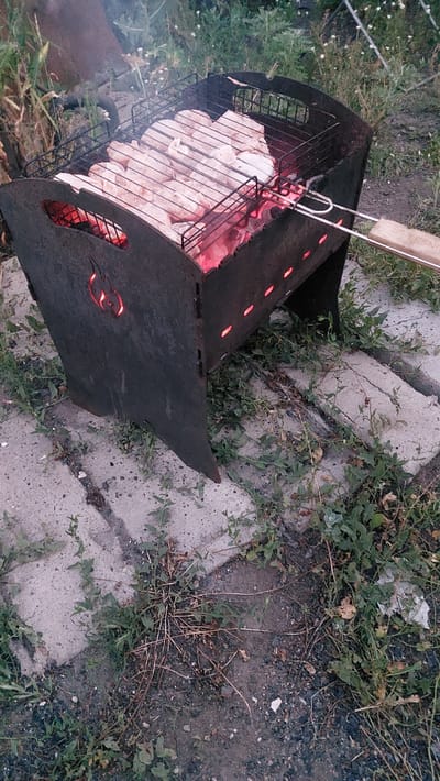 The image shows a metal barbecue grill with raw meat skewers arranged on top. The grill is fueled by glowing red coals, indicated by the visible flames and heat emanating from beneath the grate. The barbecue is situated outdoors, on a surface composed of concrete slabs and dirt, with sparse vegetation growing around it. In the background, more greenery and possibly some fencing are visible. The overall impression is that of a casual outdoor cooking setup. The location is specified as Shakhtinsk, Kazakhstan.
