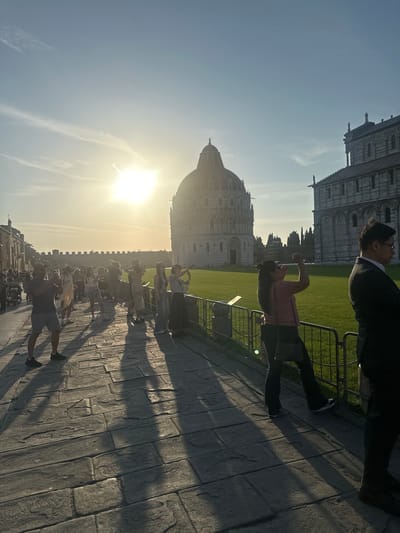 This image captures a vibrant outdoor scene in Pisa, Italy, likely during the late afternoon or early evening, given the low position of the sun. The bright sun, situated on the left side of the frame, casts a warm, golden glow and long, distinct shadows across the paved ground, creating a strong backlighting effect.

In the background, two prominent architectural marvels of Pisa are visible: the majestic Pisa Cathedral with its intricate detailing and the distinctive dome of the Pisa Baptistery, both showcasing ornate Romanesque style in light-colored stone. These buildings frame a large, well-manicured green lawn. A metal fence separates this grassy area from a wide, paved walkway that dominates the foreground and mid-ground.

The scene is bustling with numerous people, suggesting a popular tourist attraction. Many individuals are walking along the paved path, while others are standing, observing the historical landmarks. Due to the backlighting, several figures appear as silhouettes or are partially obscured. In the mid-ground on the right, a woman in a pink top and dark pants is captured in a dynamic pose, with her arms raised, possibly gesturing or posing for a photo. A man in a dark jacket stands nearby. Further in the foreground, a man in a grey shirt and shorts walks along the path. The general activity implies people enjoying a leisurely visit to a famous historical site.

The sky is clear and bright blue, with some delicate, wispy clouds visible near the sun. There is no visible text in the image.