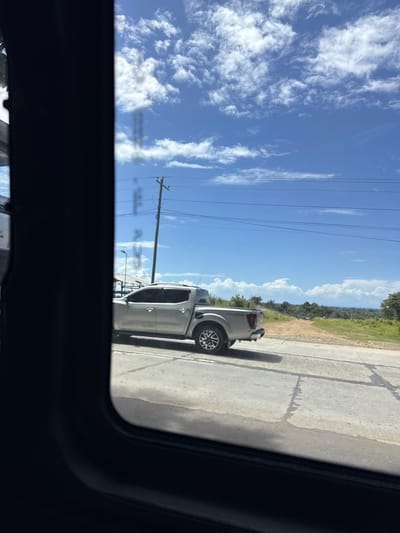 This image, taken from a vehicle's interior, shows a silver pickup truck driving on a paved road in Santa Cruz, Philippines. The truck is in the mid-ground, facing away from the viewer. The road appears to be in good condition with some water stains. Beyond the road, there is a grassy area with some trees and vegetation, leading to a distant view of the sea or a large body of water under a bright blue sky with scattered white clouds. Electrical poles and wires are visible in the background. The foreground is dominated by the dark interior of the vehicle the photo was taken from, specifically the window frame and part of the dashboard. There are no people clearly visible in the scene.