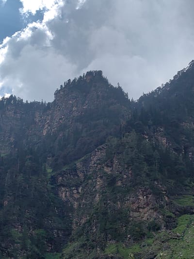 This is a vertical, eye-level, full shot of a steep, tree-covered mountain range under a cloudy sky. The mountain slopes are rocky and rugged, with dark green pine trees densely covering much of the terrain. The clouds are a mix of white and dark grey, suggesting an overcast or partially sunny day. In the upper left corner, there is a patch of bright blue sky. The image appears to be taken from a lower elevation, looking up at the imposing mountain face. The location is Kothi, India, a region known for its mountainous landscape. There are no people or animals visible in the image.