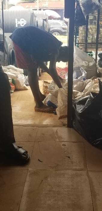 The image depicts an indoor space transitioning to an outdoor environment in Ado-Ekiti, Nigeria. The interior floor consists of light brown square tiles, exhibiting dark stains and smudges. A person with dark skin, wearing a dark short-sleeved top and red shorts, is prominently positioned in the center, bent over at the waist. This individual wears brown flip-flops and has their right arm extended into an open white woven sack on the floor. Behind this person's legs, a light brown wicker basket is present. To the right of the central figure, a collection of white woven sacks and one large black plastic sack are stacked against a dark vertical metal beam, which defines the right edge of the immediate indoor area. In the extreme foreground on the left, the lower leg and foot, clad in a dark sandal, of a second person are visible.

Through the doorway, the outdoor scene includes two large, dark grey cylindrical plastic water storage tanks immediately outside the opening on the left, one displaying a white logo. To the right of these tanks, a dark-colored SUV is parked on an unpaved surface. The background reveals corrugated metal rooftops of distant structures, interspersed with green foliage. A white or light-colored umbrella or canopy is partially visible in the far-right background. The overall lighting suggests daytime.