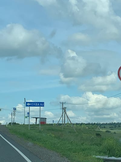 A road sign in the city of Суда, Russia, indicates a distance of 0.1 kilometers in a direction to the left. The sign is blue with white text and an arrow pointing left. In the background, there is a green grassy field with trees and a cloudy sky. Several other signs, including a parking sign and a speed limit sign, are visible near the main road sign. There are also power lines and a small bus stop structure in the distance. The perspective suggests the photo was taken from a vehicle driving on the road.