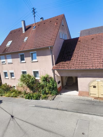 The image shows a residential street in Hardthausen am Kocher, Germany, on a clear, sunny day. A two-story building with a red tiled roof dominates the center and right side of the frame. The building has a light pink facade with multiple windows, some featuring white rolling shutters. A smaller, darker-roofed building is visible behind the main structure, partially obscured.
In front of the main building, a driveway leads to an arched opening, revealing a set of concrete stairs ascending into the building. To the right of the opening, there is a large, light-colored double wooden door with black hardware. Lush green foliage and flowering bushes are planted along the base of the building, extending towards the street.
The street in the foreground is paved with asphalt and has a raised concrete curb. The shadow of a utility pole and wires stretches across the road, indicating the presence of electrical infrastructure. The sky is a bright, clear blue. No people or vehicles are visible in the image.