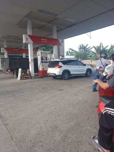 A white SUV is parked at a Pertamina gas station in Cibarusah, Indonesia, with its rear end positioned over a gas pump. To the left of the SUV, a black truck is parked. A person on a motorcycle, wearing a helmet, is standing to the right of the SUV. Another person is visible on a red motorcycle in the foreground, partially obscuring the view. The gas station has red and white signage, with "3 PERTALITE BIOSOLAR" visible on one sign. The ground is paved with asphalt and concrete. Trees and foliage are visible in the background.
