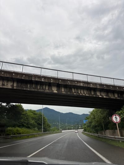 The image depicts a highway in Chakvi, Georgia, passing under a concrete overpass. The sky is overcast and gray. The road is wet, indicating recent rain, and marked with white lines. A white car is visible in the distance on the highway. On the right side of the road, there is a circular speed limit sign with "5 M" inside a red border, indicating a 5-meter height restriction. Lush green trees and mountains form the backdrop. The overpass has a metal railing on top and appears weathered.