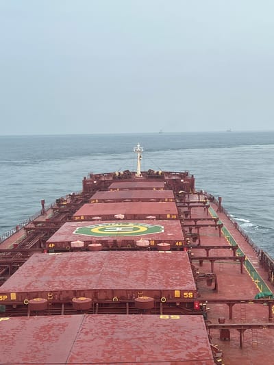 The image shows the deck of a large cargo ship sailing on a body of water. The deck is covered in red hatches, some of which have yellow markings indicating their identification numbers, such as "4P," "5S," and "6S." A helideck with a green circle and yellow markings is visible on one of the hatches. The sea is calm with a slight chop, and the sky is overcast and grey. In the distance, a few other ships can be seen on the horizon. The perspective is from the stern of the ship, looking forward. The scene appears to be at sea, not in a specific city location in Malaysia.