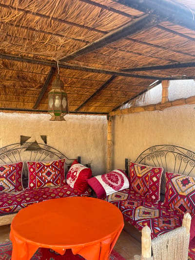 A view from beneath a thatched roof with black beams. A decorative lantern hangs from the roof. The scene transitions to a beige wall with a decorative metal shelf displaying patterned pillows in red, white, and gold. An orange table is visible in the foreground. The image suggests an indoor space with a rustic, possibly Middle Eastern or African-inspired design.