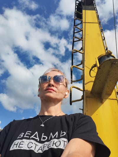 A low-angle medium shot shows a woman looking up at the sky, with a yellow crane structure dominating the right side of the frame. The woman, with blonde hair and sunglasses reflecting the clouds, wears a black t-shirt with silver Cyrillic text that reads "ВЕДЬМЫ НЕ СТАРЕЮТ" (Witches don't age). She has small white wireless earbuds in her ears and a delicate silver necklace with a pendant. The background is a bright blue sky with scattered white clouds. The yellow crane, likely industrial equipment, appears weathered with visible rust on its ladder rungs. The image was taken outdoors in daylight. The scene is situated in Krokhino, Russia, a location known for its industrial heritage and the abandoned Church of the Nativity of the Blessed Virgin Mary, which is partially submerged in the Sheksna River.
