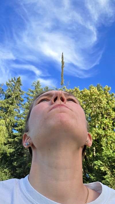 A young Caucasian woman is looking upwards with a blade of grass balanced vertically on her nose. She is wearing a white t-shirt and small earrings. The background is a bright blue sky with wispy white clouds, and a dense green forest with tall trees. The image is taken from a low angle, emphasizing the sky and the woman's face. The location is not specified in the image.