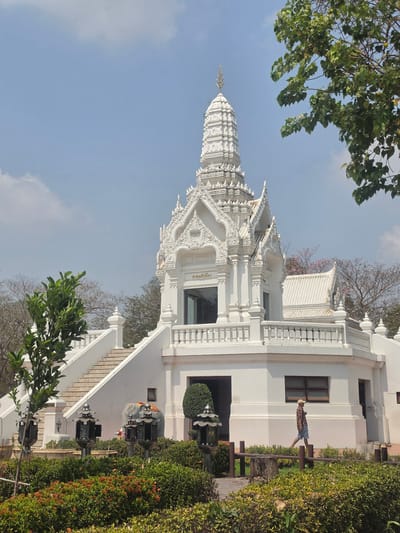 This image depicts Wat Ratchaburana, a Buddhist temple in the Historic City of Ayutthaya, Thailand. The temple is predominantly white, featuring intricate architectural details. A person stands on a bench, possibly observing the scene, next to the temple structure. Green vegetation borders the building, and the sky is partly cloudy.
