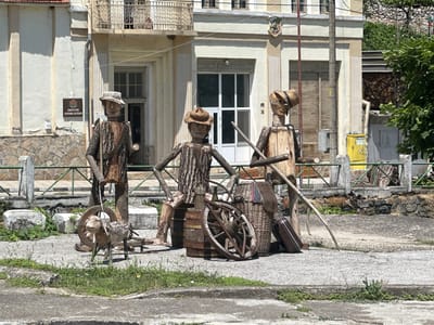 In Golyamo Belovo, Bulgaria, a series of wooden sculptures depicting figures in rustic attire stand in front of a building with a sign displaying Bulgarian text, likely indicating a municipal building. The sculptures, made from rough-hewn logs and branches, are positioned around an old wooden cartwheel and barrels. One sculpture is a roughly carved sheep with a wooden wheel behind it. The building's facade is light-colored, with a balcony on the upper floor. The ground in front of the building is paved, with patches of grass and weeds growing through cracks. A green metal fence runs along the property line. The overall scene suggests an outdoor display or a decorative arrangement in a public space.