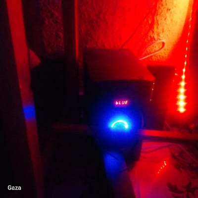 The scene is an interior space located in Kamera ward, Kenya, characterized by low light conditions dominated by red and blue illumination. Centrally positioned is a dark electronic device, likely an audio system, exhibiting a red LED display that reads "BLUE" and a prominent blue circular light with its lower arc illuminated. To the immediate right, a strip of vertical red LED lights is visible against a rough-textured wall, which is also bathed in red light. White cables are seen near the top right, connected to components obscured by the intense red lighting. On the left side, a vertical dark reddish-brown structure or surface reflects a small blue light. The foreground shows a faintly patterned surface, possibly a tabletop. The text "Gaza" is present in white at the bottom left corner of the image. No individuals are present.