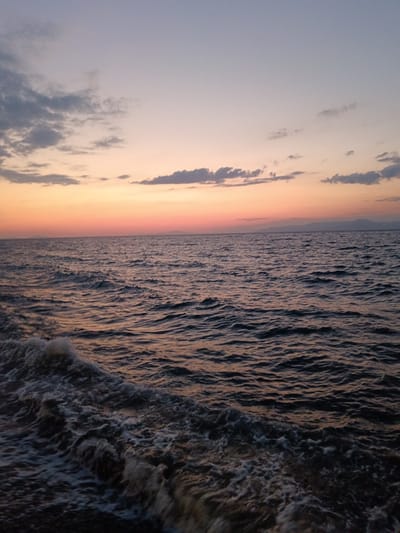 The image depicts a body of water, likely the sea, with waves crashing in the foreground. The water is dark and choppy, reflecting the colors of the sky. The sky above is a gradient of colors, transitioning from a pale blue at the top to hues of orange and pink near the horizon, indicating either sunrise or sunset. Scattered clouds are visible in the sky, some appearing darker and more defined than others. In the distance, a faint landmass or hills can be seen on the horizon. The scene appears to be natural and serene.
The location is the Municipal Unit of Vrachneika, Greece. This is a coastal area known for its natural beauty and proximity to the sea. The specific scene captured in the image is likely a view from the coastline or a boat at sea, showcasing the natural environment of Vrachneika during the twilight hours. There are no people or man-made structures prominently visible in the image, emphasizing the natural elements of the seascape.