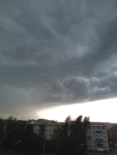 The image depicts a street view in Kyshtym, Russia, under a dramatic sky filled with dark, heavy storm clouds. The foreground is dominated by dense, dark green trees, with some of their branches appearing to be swept by wind, suggesting gusty conditions. A lamppost is visible on the left side. Beyond the trees, several multi-story apartment buildings, typical of Soviet-era architecture, are lined up horizontally across the frame. These buildings are gray and show various window configurations and balconies. In the distance, a lighter, almost hazy patch of sky suggests the presence of rain or a distant storm cell. The overall atmosphere is somber and overcast, indicative of impending or ongoing bad weather.