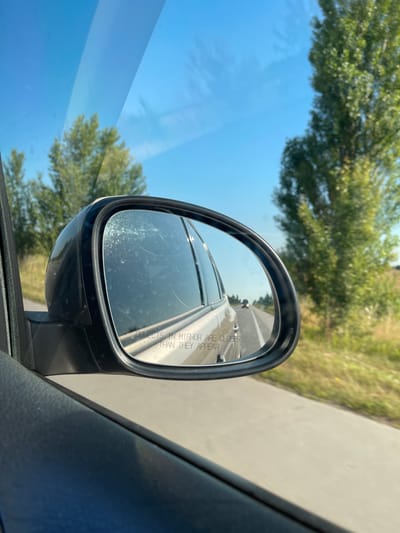 The image shows a view from inside a car, looking out the side window and into the side mirror. The side mirror reflects a road with other vehicles driving on it, and trees lining the side of the road. The sky is blue and clear, indicating it is daytime. The car appears to be in motion, as suggested by the blurred trees in the background. The mirror has the text "OBJECTS IN MIRROR ARE CLOSER THAN THEY APPEAR" etched into it. The interior of the car, including the door panel and window frame, is visible in the foreground. There is no information in the image that indicates the location of Riznya, Ukraine.