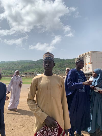 The image shows a group of people in what appears to be the outskirts of Naraguta Hausawa, Nigeria. The foreground features several individuals in traditional Hausa clothing, including a man in a tan robe and a patterned cap prominently facing the camera. To his right, partially visible, is someone in a dark blue garment with patterned embroidery. Further back, a woman in a blue hijab is holding a smartphone. A woman in a light-colored garment walks behind the group.
In the background, a truck is parked. Beyond the truck, the terrain slopes up to a hill covered in vegetation. An unfinished building can be seen on the hillside. The ground is dusty. The sky above is cloudy.