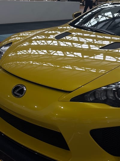 The image shows the front of a yellow Lexus LFA supercar on display at the Lexus showroom in Nagoya, Japan. The car is positioned indoors, with a reflective ceiling overhead that casts a grid-like pattern onto the car's hood. The showroom appears spacious, with a dark floor and a lighter wall visible in the background. A person, likely an employee or visitor, is partially visible in the upper right portion of the image, their silhouette reflected on the car's windshield. The car's design features prominent headlights, a distinctive Lexus emblem on the hood, and aggressive vents. The lighting is bright, emphasizing the car's metallic paint finish.