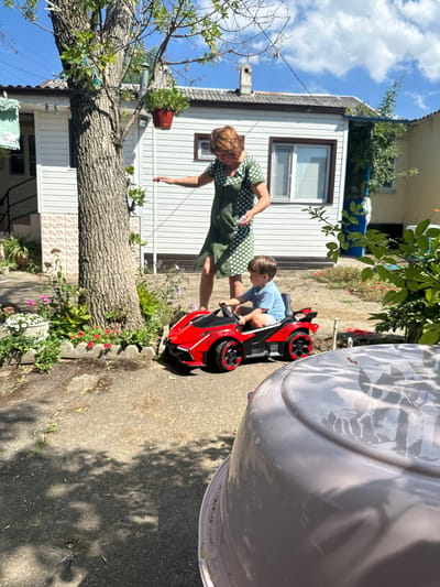 In the city of Temryuk, Russia, on a sunny day with scattered clouds, a woman in a green polka-dot dress is standing in a yard. She is reaching out with her left hand towards a young child sitting in a red toy car. The child, wearing a blue t-shirt and grey shorts, is holding onto the steering wheel. The toy car has "SW98" written on its side. Behind them, a white house with a dark roof and windows is visible. A large tree stands to the left of the woman and child, with a potted plant hanging from one of its branches. In the foreground, a pink object, possibly a large basin or tub, takes up the bottom portion of the image, casting shadows on the asphalt ground. Flowers are planted in front of the house and around the base of the tree. The overall scene depicts a playful moment between a woman and a child in a residential outdoor setting.