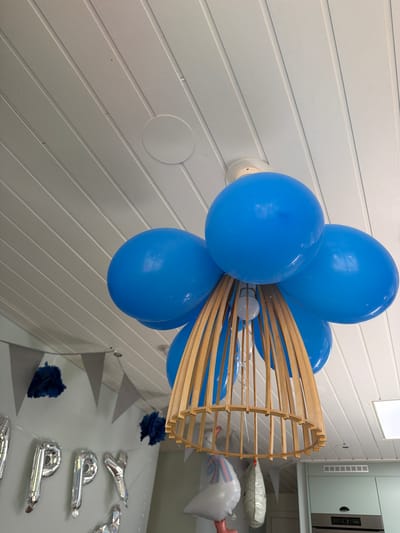 The image shows a close-up, low-angle view of a ceiling decorated with blue balloons and a wooden light fixture. The ceiling is made of white-painted wooden planks. A circular white vent or fixture is visible on the ceiling. The light fixture, made of thin, curved wooden slats, is suspended from the ceiling and appears to be a lampshade. Several bright blue balloons are clustered around the top of the light fixture, some attached to it and others floating above. Below the balloons and light fixture, a banner of grey triangular flags is hung, along with dark blue tissue paper pom-poms. To the left, silver foil balloons spelling out "HAPPY" are partially visible. Further to the right, other decorations like a stork-shaped balloon are present, as well as a mint green cabinet with a stainless steel appliance. The overall scene suggests a celebratory event, possibly a baby shower, taking place indoors. There are no people visible in the image. The location is in Heijen, Netherlands.